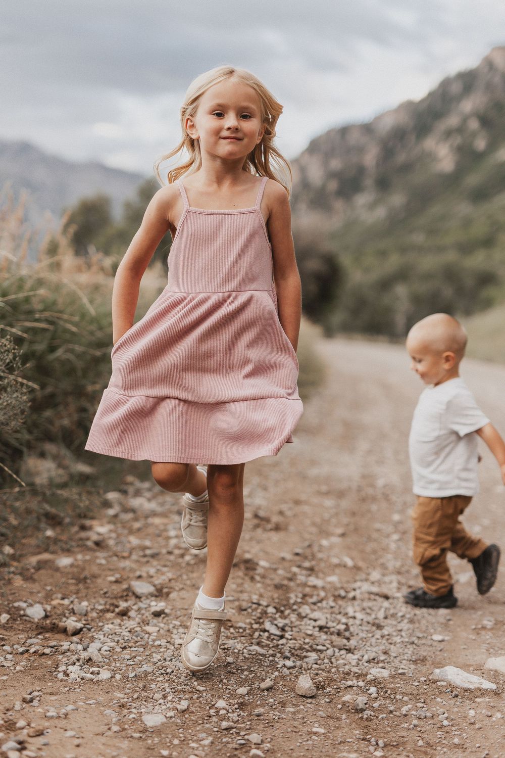 A young girl in a pink dress skips on a mountain dirt path while a toddler boy walks behind her.