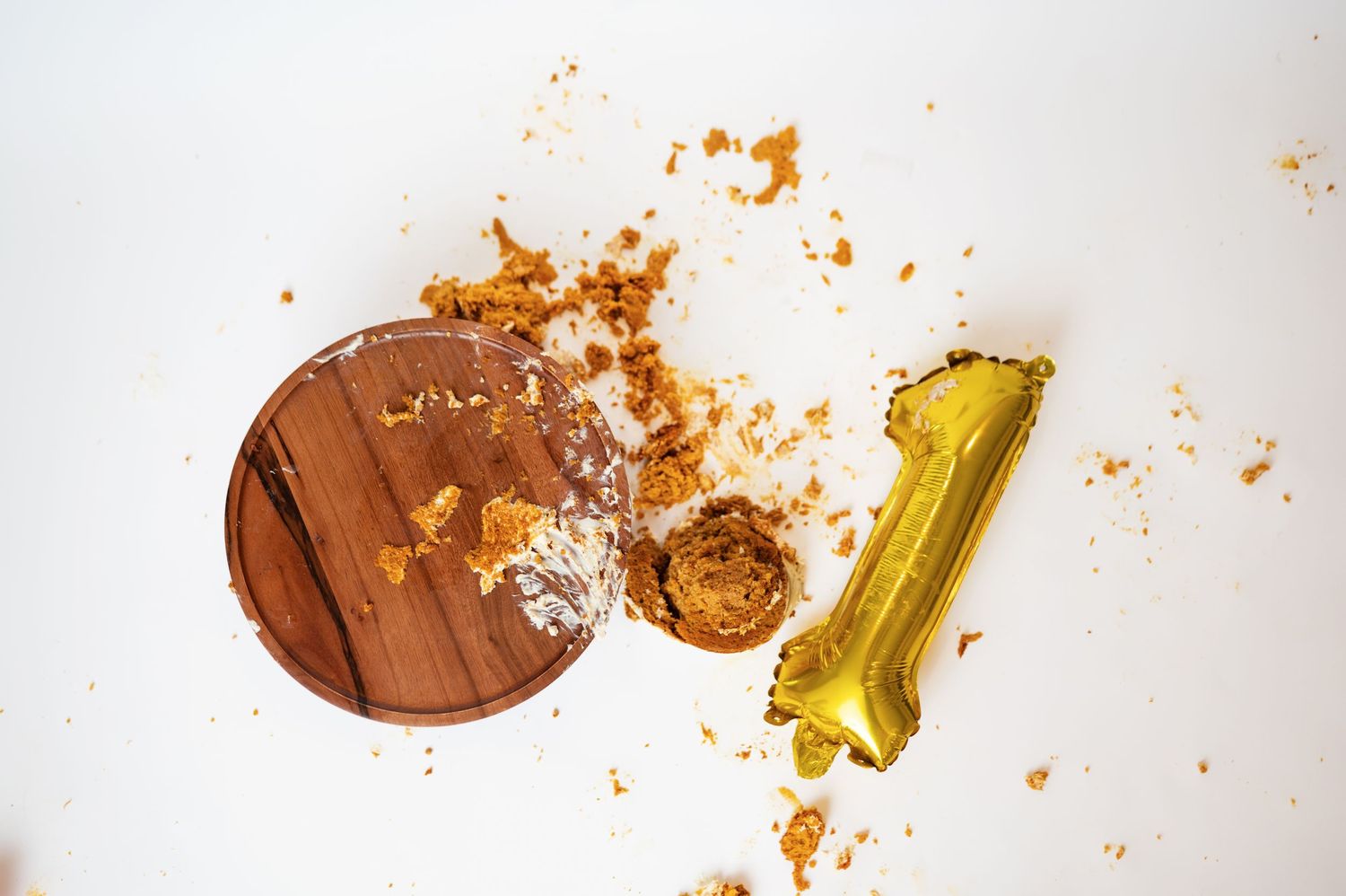 aerial view of aftermath of cake smash session in home; gold foil "1" balloon, cake stand with icing and cake remanants, cake upside down