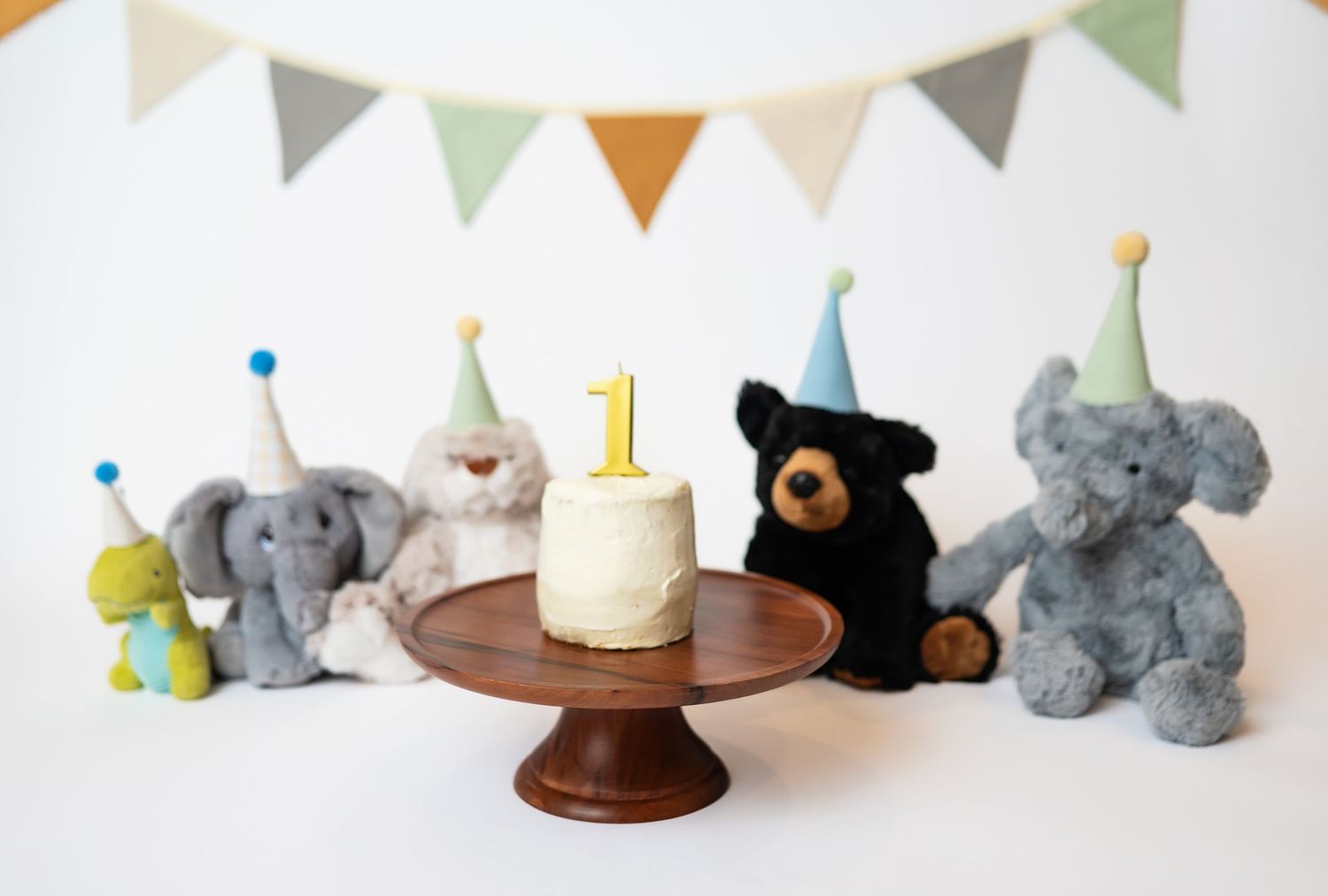 vanilla iced cake on wooden cake stand surrounded by stuffed animals with party hats on; the future scent of a  1 year cake smash session