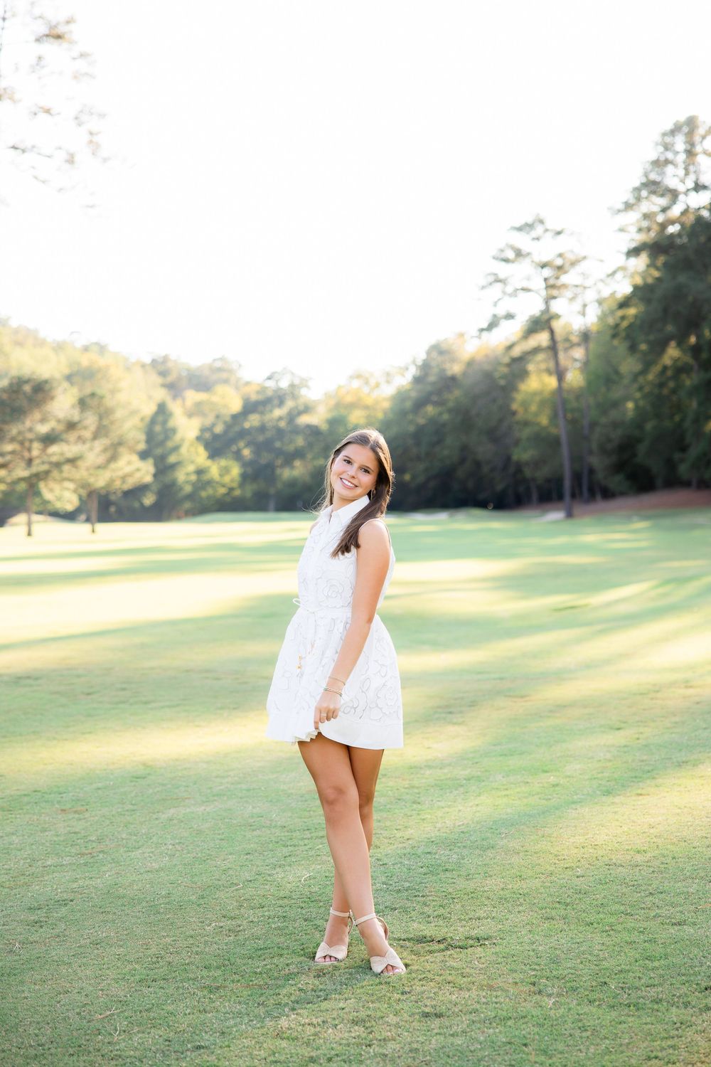 Brunette woman in white sleeveless dress stands smiling on sunny green golf course lawn.