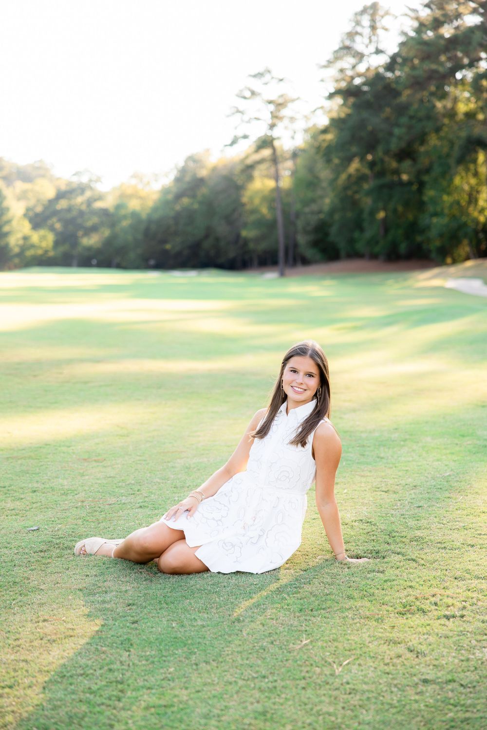 Smiling brunette woman in white dress sits on lush green golf course grass at sunset.