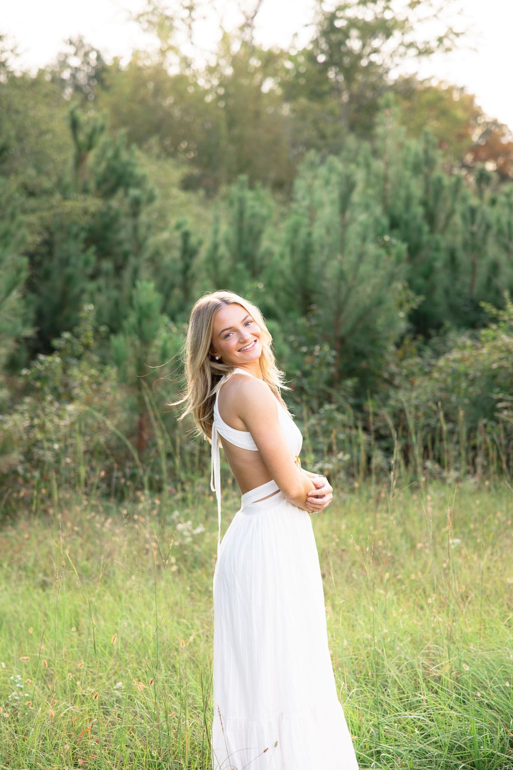 Smiling blonde woman in white cutout dress poses in green meadow with pine trees behind her.