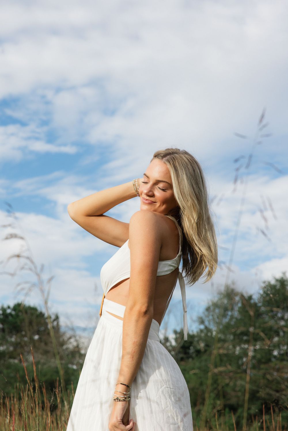 Blonde woman in white cutout dress tilts head back in sunlit field under partly cloudy sky.