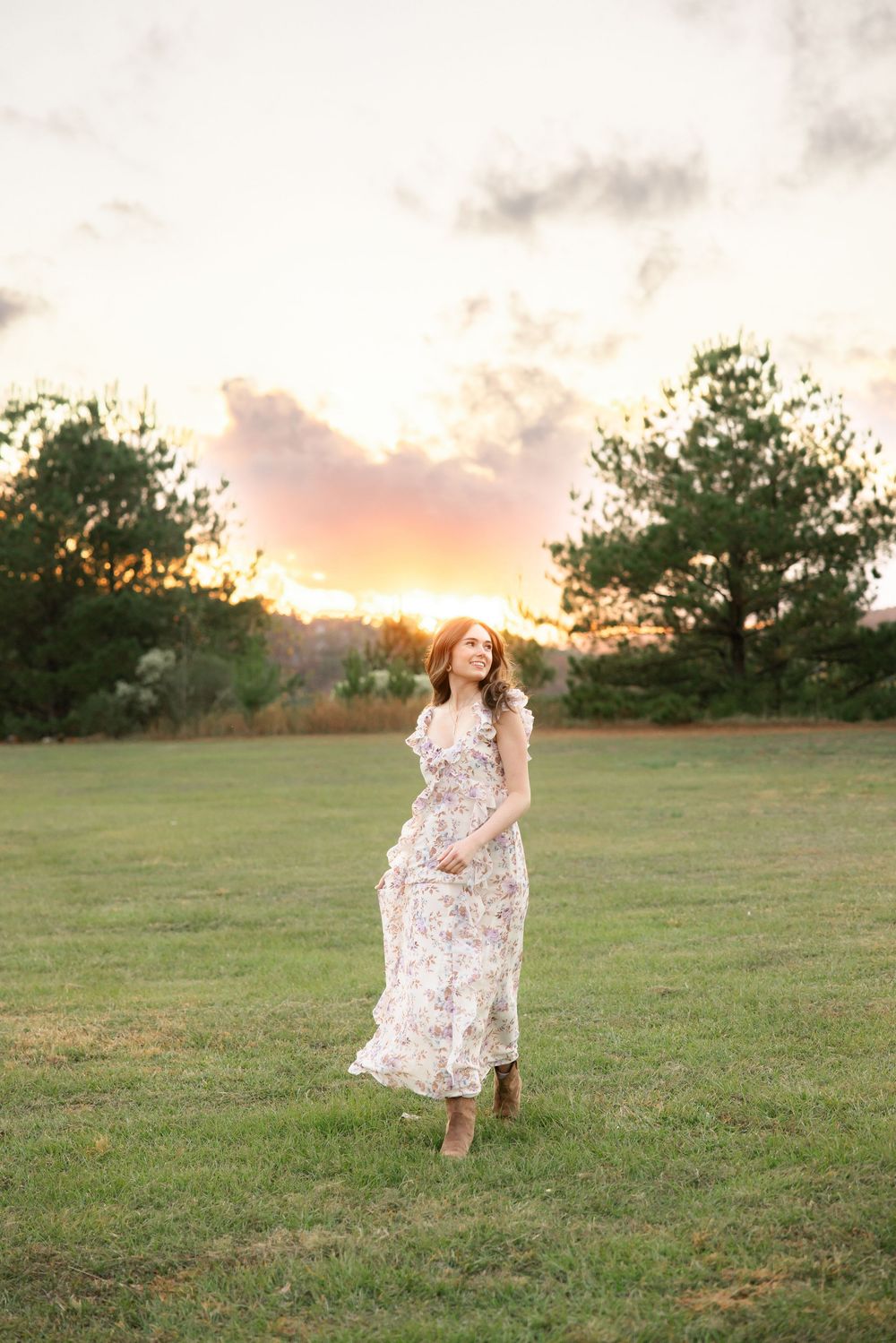 Young woman in floral maxi dress and cowboy boots twirls in open field during golden sunset hour.