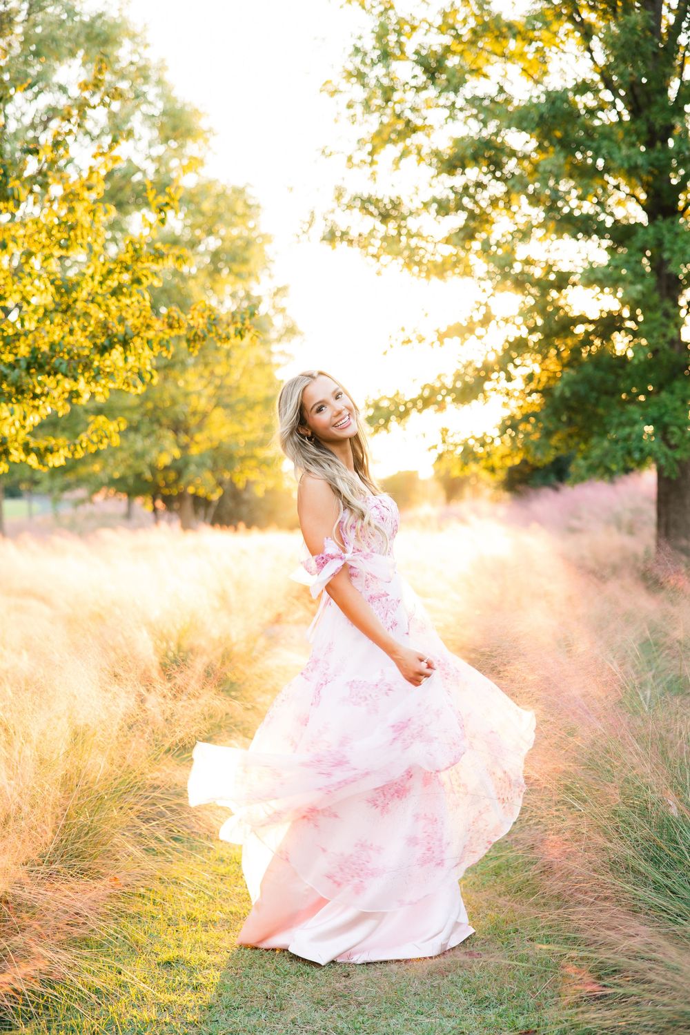 Blonde woman twirls in pink floral gown on path through tall grass at golden hour.