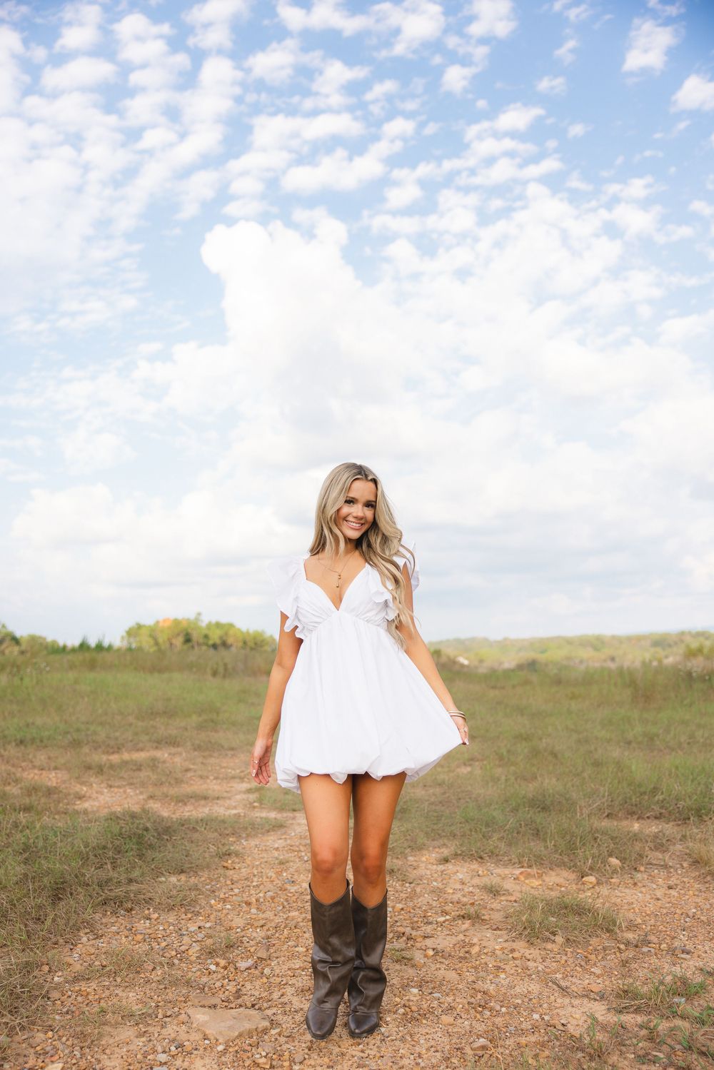 Smiling blonde woman in white mini dress and cowboy boots stands in open field under cloudy sky.