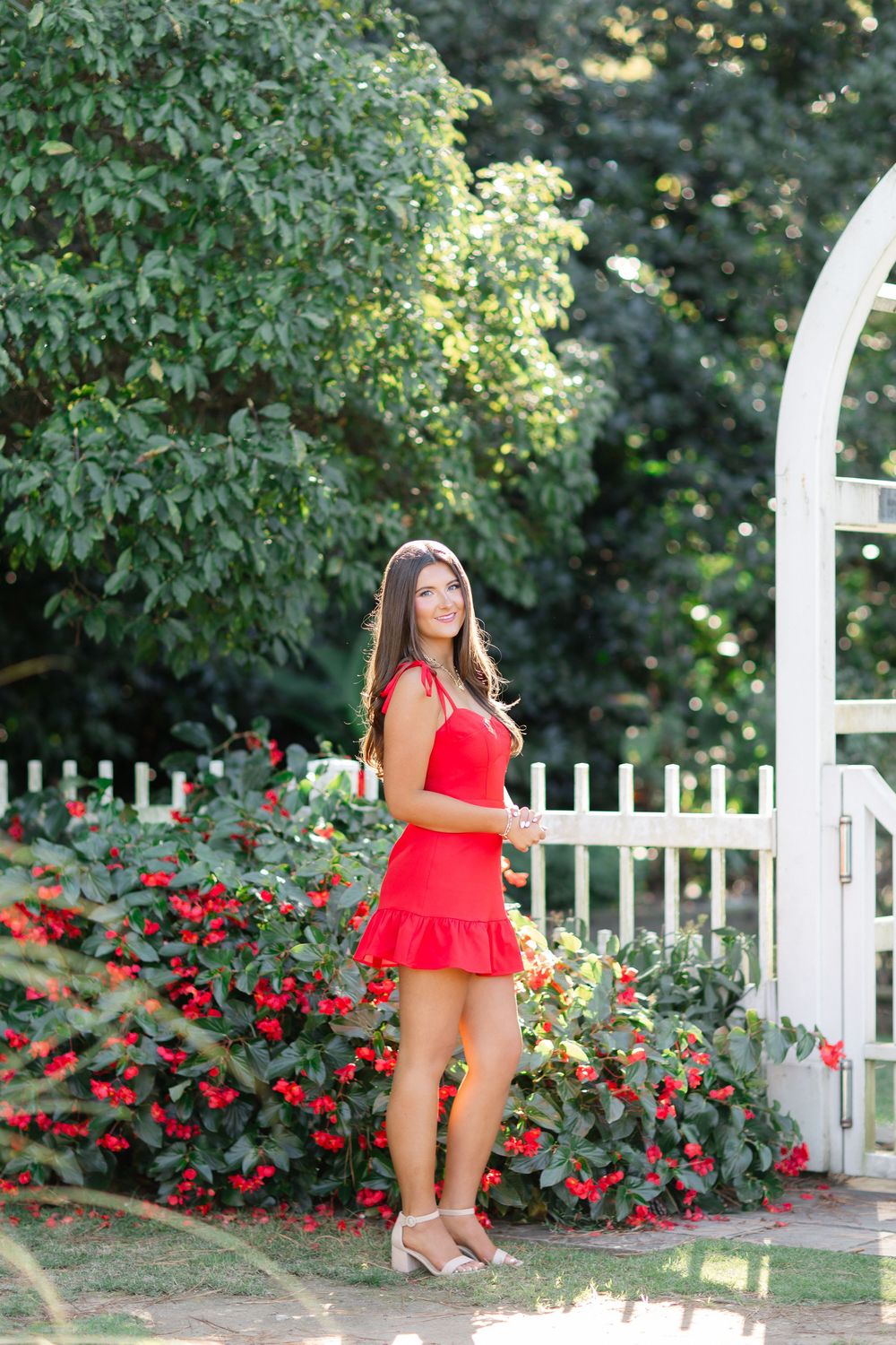 Young woman in red mini dress poses by white garden gate with red flowers and lush greenery.