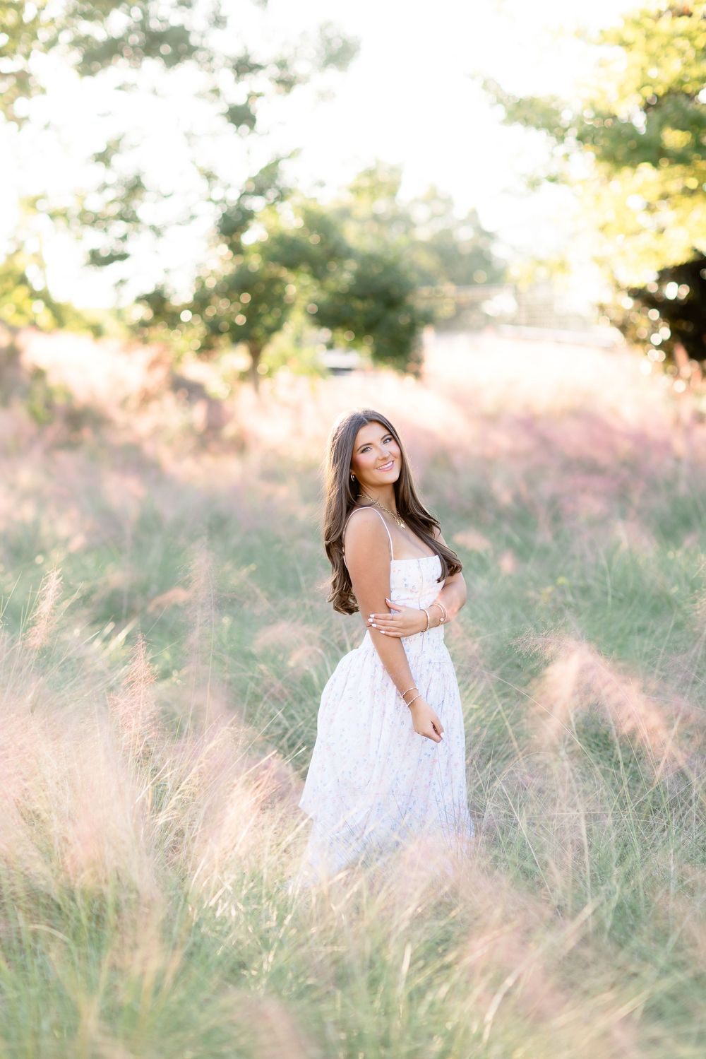Brunette woman in white lace dress stands in golden field of tall grass at sunset.