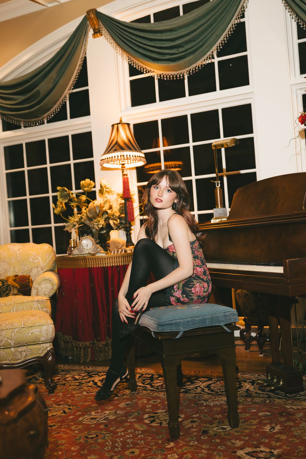 Young woman in floral dress and black tights poses on piano bench in grand traditionally decorated room.