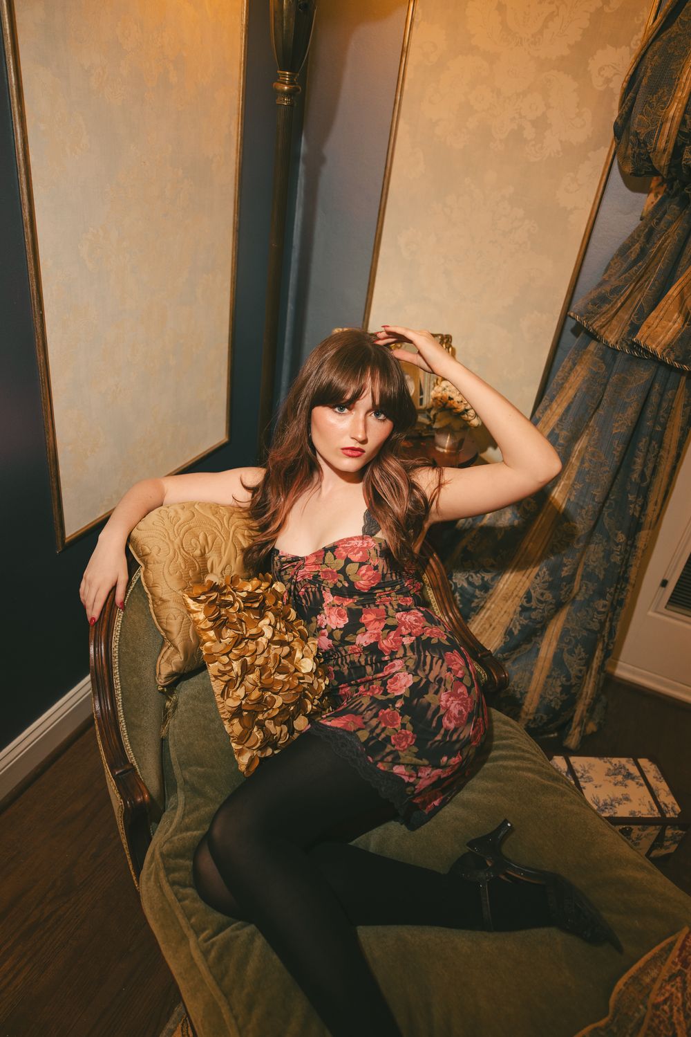 Brunette woman in floral dress and black tights poses in velvet armchair in vintage-styled elegant room.