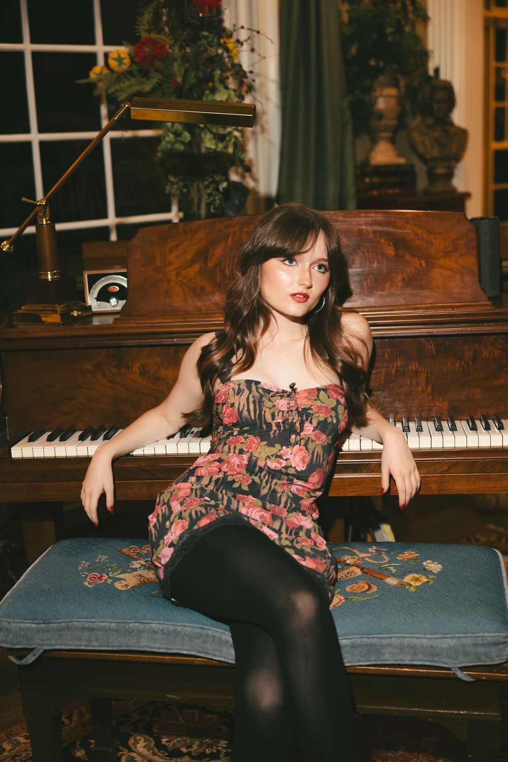 Brunette woman in floral dress sits at vintage wooden piano in elegantly decorated traditional living room.