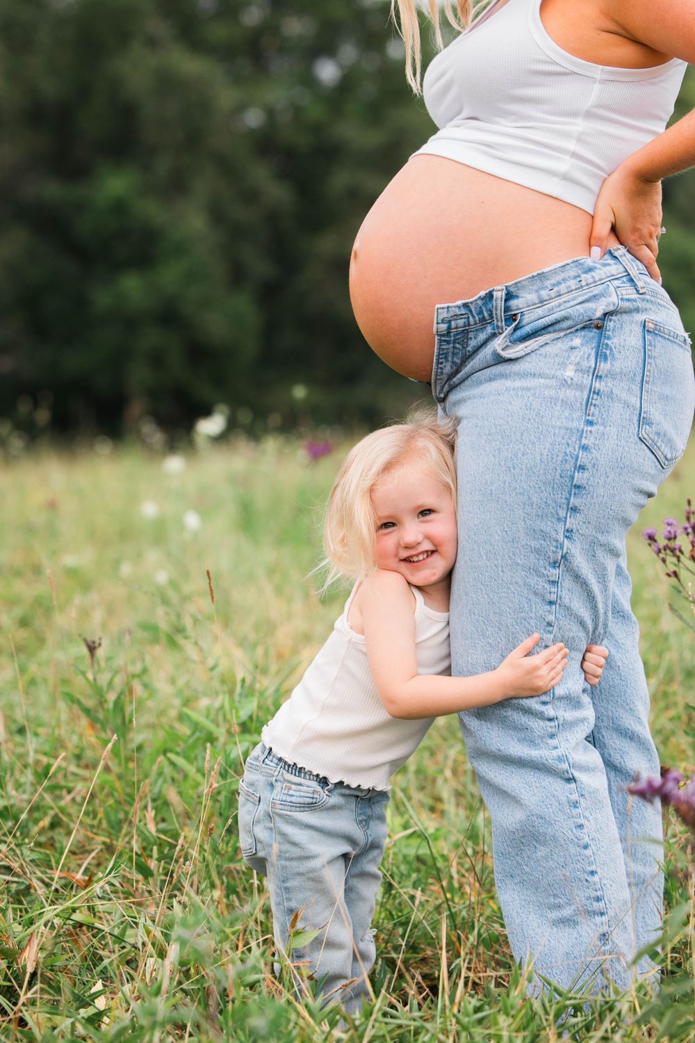 Toddler hugs pregnant mother's leg in sunny meadow with wildflowers.