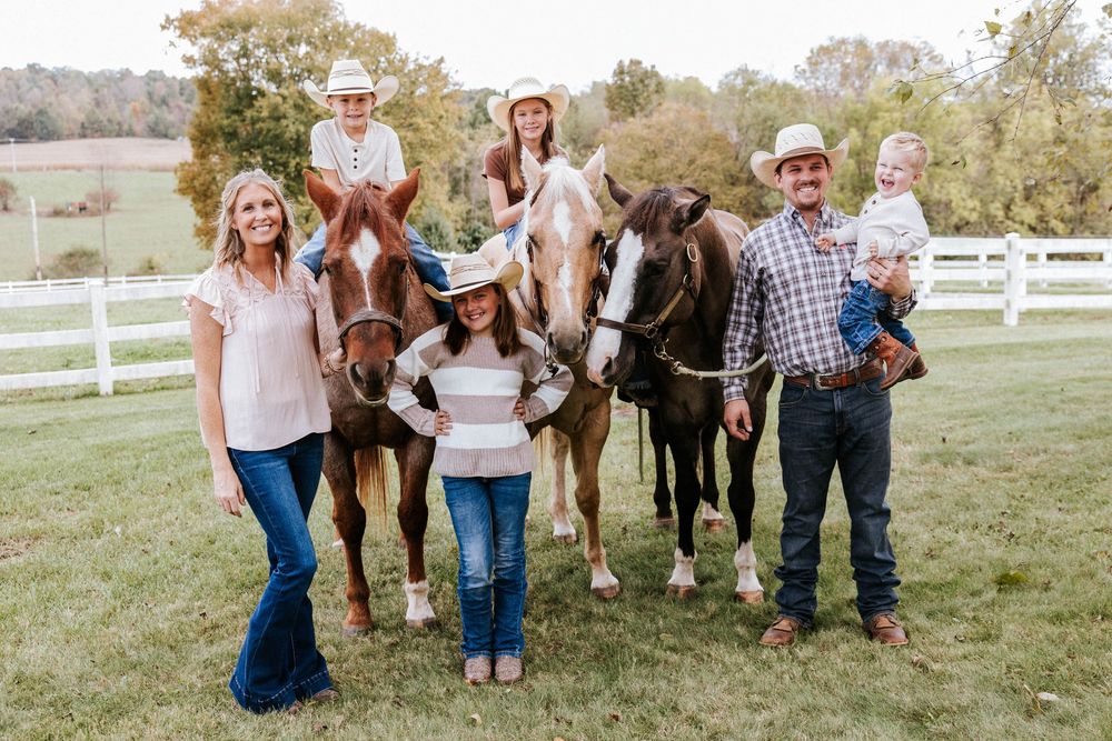 Family poses with their horses on a scenic farm with white fences and autumn trees in the background.