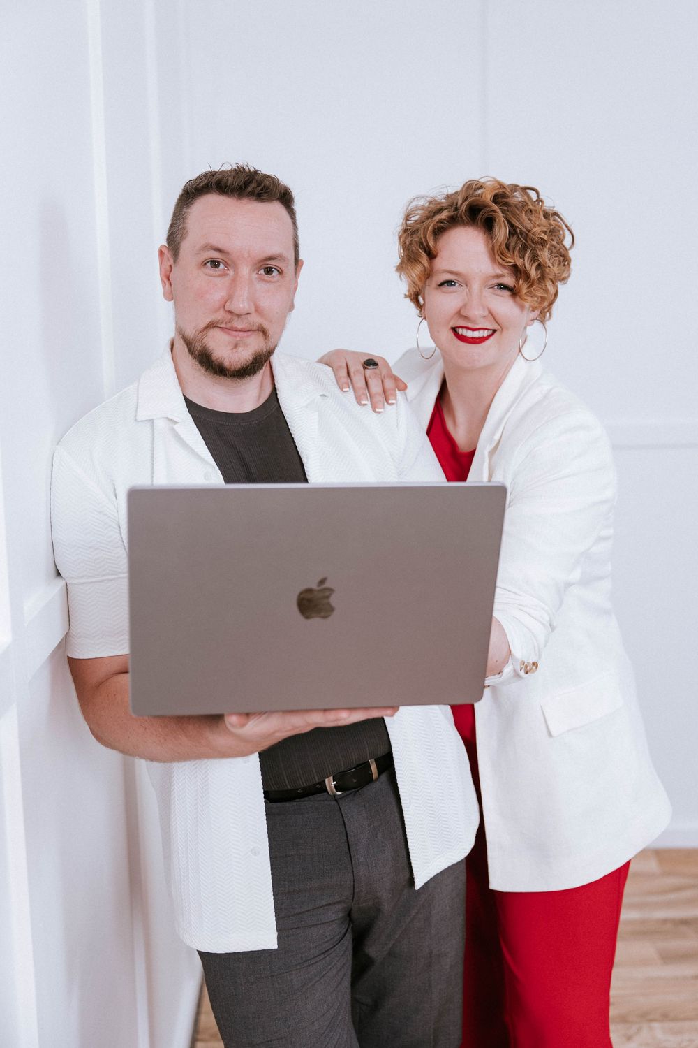 Two people in contrasting outfits pose with a laptop in a bright white room.