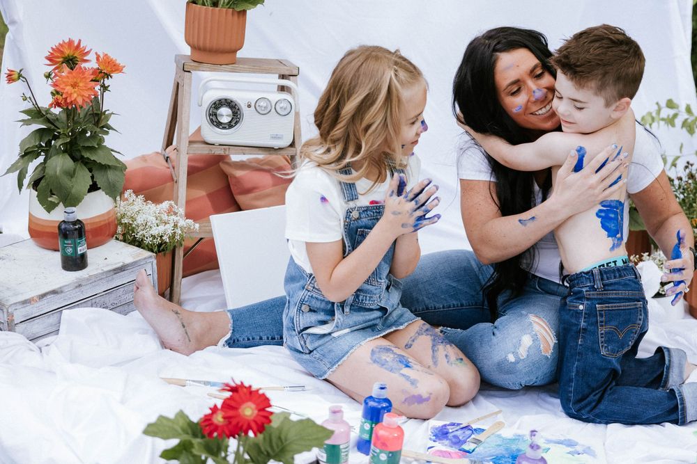 A mother and two young children laugh and play with colorful paint in a bright, flower-decorated studio setting.
