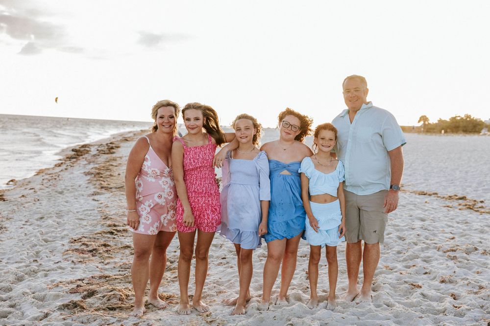 A group of people wearing casual summer attire pose together on a sandy beach at sunset.