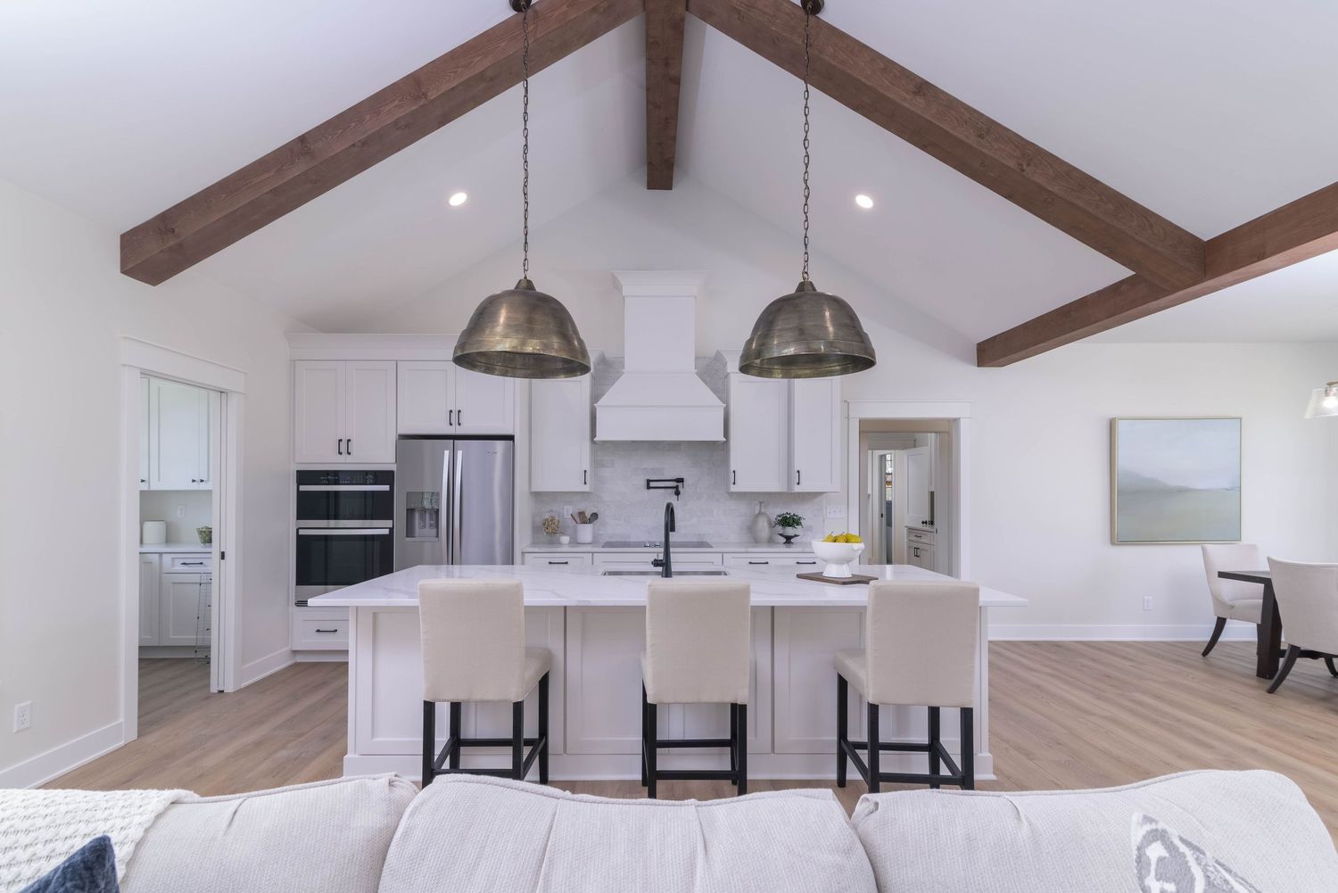Modern white kitchen with vaulted ceiling, exposed wooden beams, large island, and industrial pendant lights.
