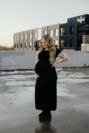 A pregnant figure in a flowing black dress stands in an urban setting at sunset.