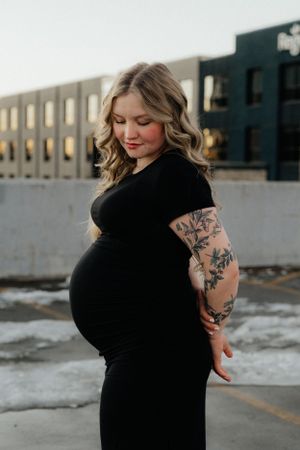 A pregnant person in a black dress with tattoo sleeves poses outdoors against an industrial building backdrop.