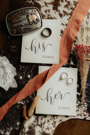 Wedding vow books labeled 'his' and 'her' with rings and western belt buckle on cowhide background.