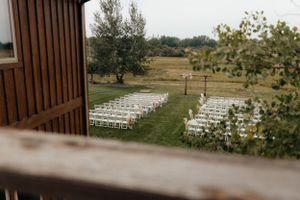 White ceremony chairs set up on a lawn viewed through a wooden window frame.