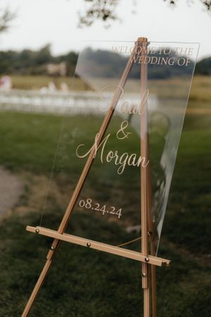 A clear acrylic wedding welcome sign on wooden easel displaying wedding details.