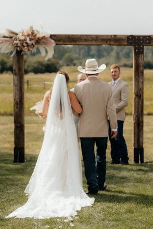A rustic outdoor wedding ceremony under a wooden arch decorated with dried flowers and pampas grass.