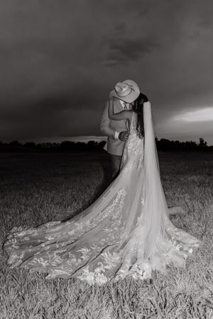 A romantic black and white photo of a couple embracing in a field with the bride wearing a long flowing lace wedding dress.
