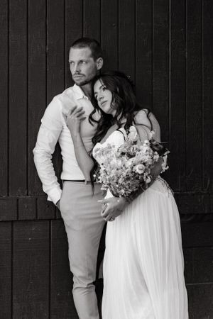 Black and white wedding portrait of a couple posing against a wooden wall with a floral bouquet.