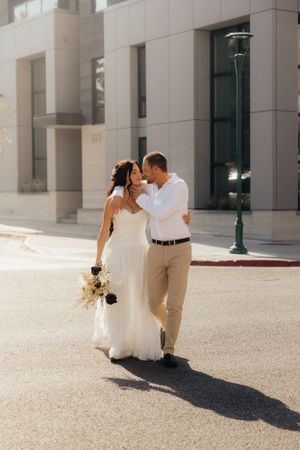 Newlyweds walking together on a sunny day in an urban setting with the bride holding a bouquet.