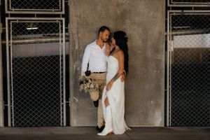 Couple in wedding attire embracing against an industrial concrete wall with chain link fencing.
