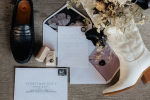 Wedding details flatlay with shoes, invitation, and dried flowers on wooden surface.