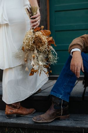 Close-up of brown boots and a white dress with dried flower bouquet on wooden steps.