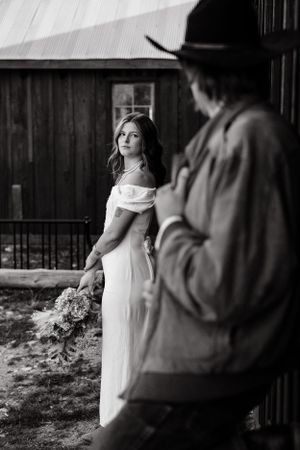 Black and white photo of bride with bouquet near rustic barn building.