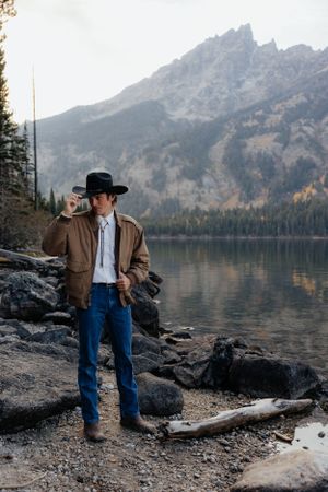 Groom in western wear poses by mountain lake during golden hour.