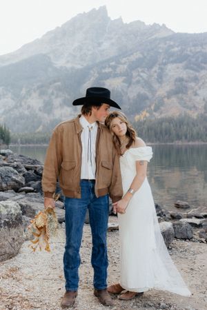 Western-styled couple stands together by mountain lake holding small teddy bear.