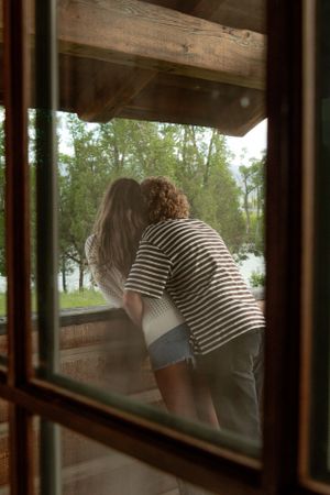 A couple leans out of a rustic cabin window overlooking a lake and forest.