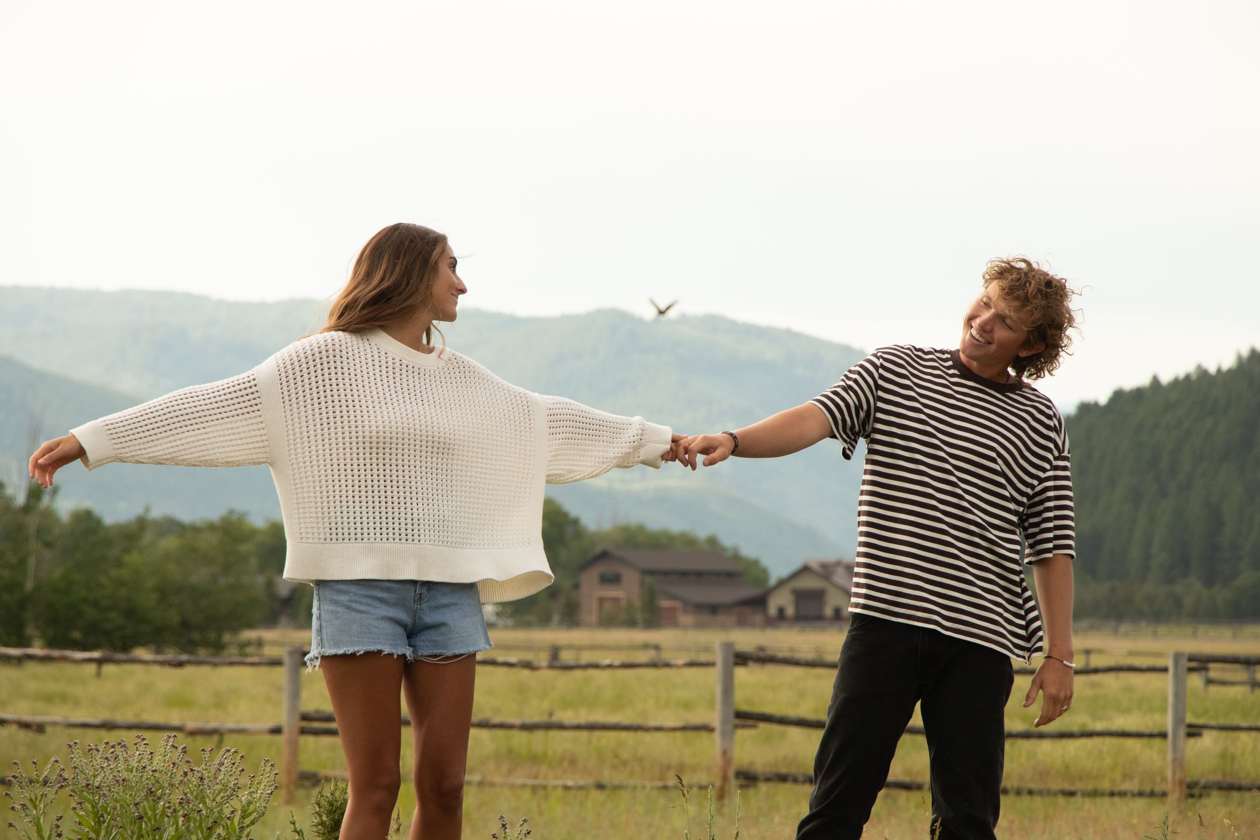Two people hold hands and dance in a mountain meadow with wooden fencing.
