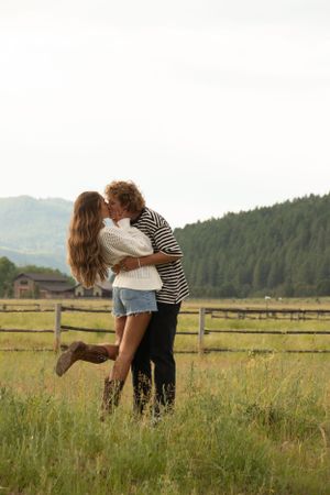 A romantic embrace in a mountain meadow with rustic fence and forest backdrop.