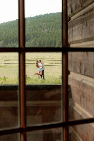 View through a cabin window of a couple embracing in a mountain meadow.