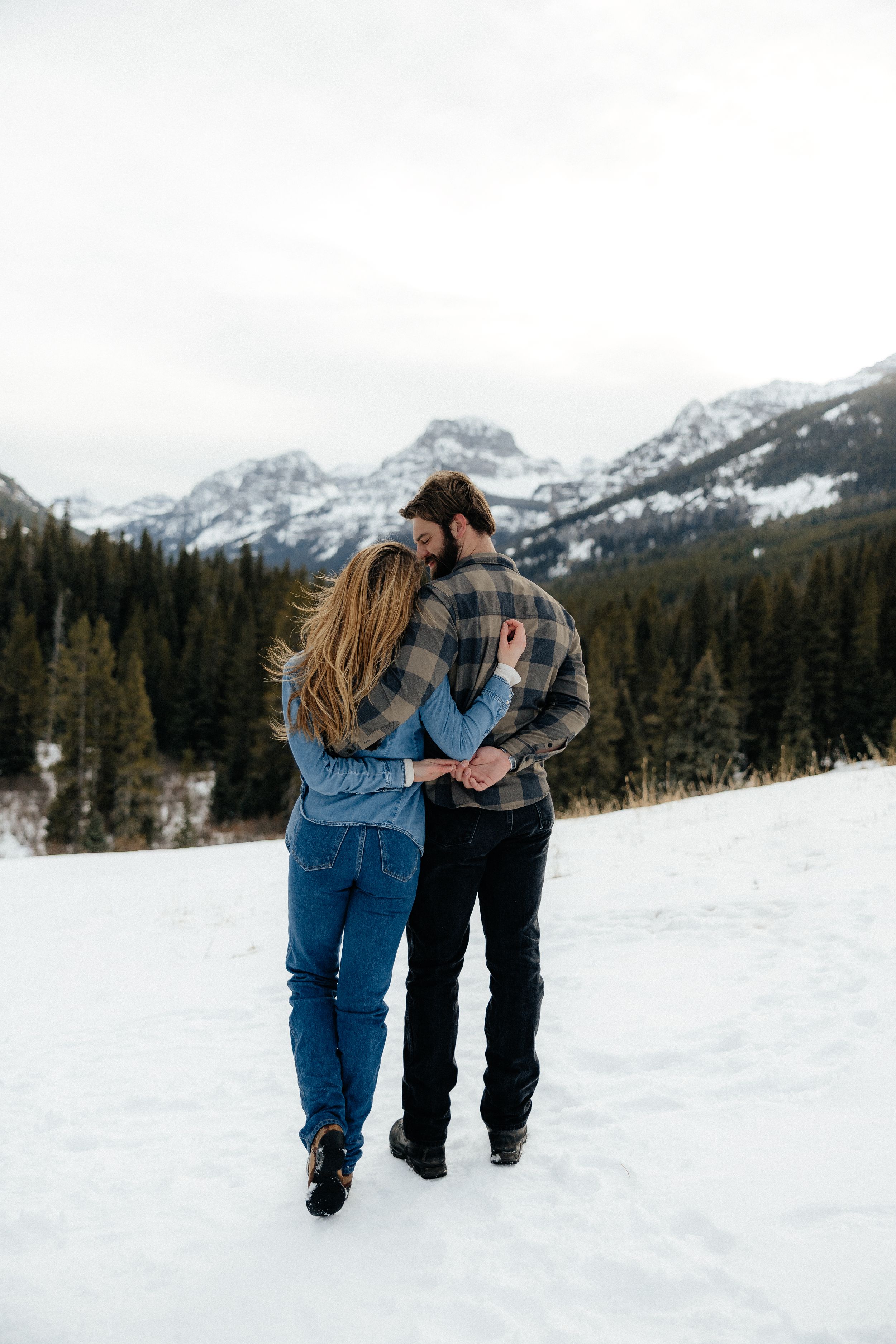 A couple walks together through snowy mountains and pine forests in winter, embracing against a scenic mountain backdrop.