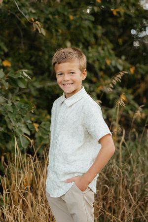 Young child in white button-down shirt and khaki pants poses outdoors against natural foliage background.