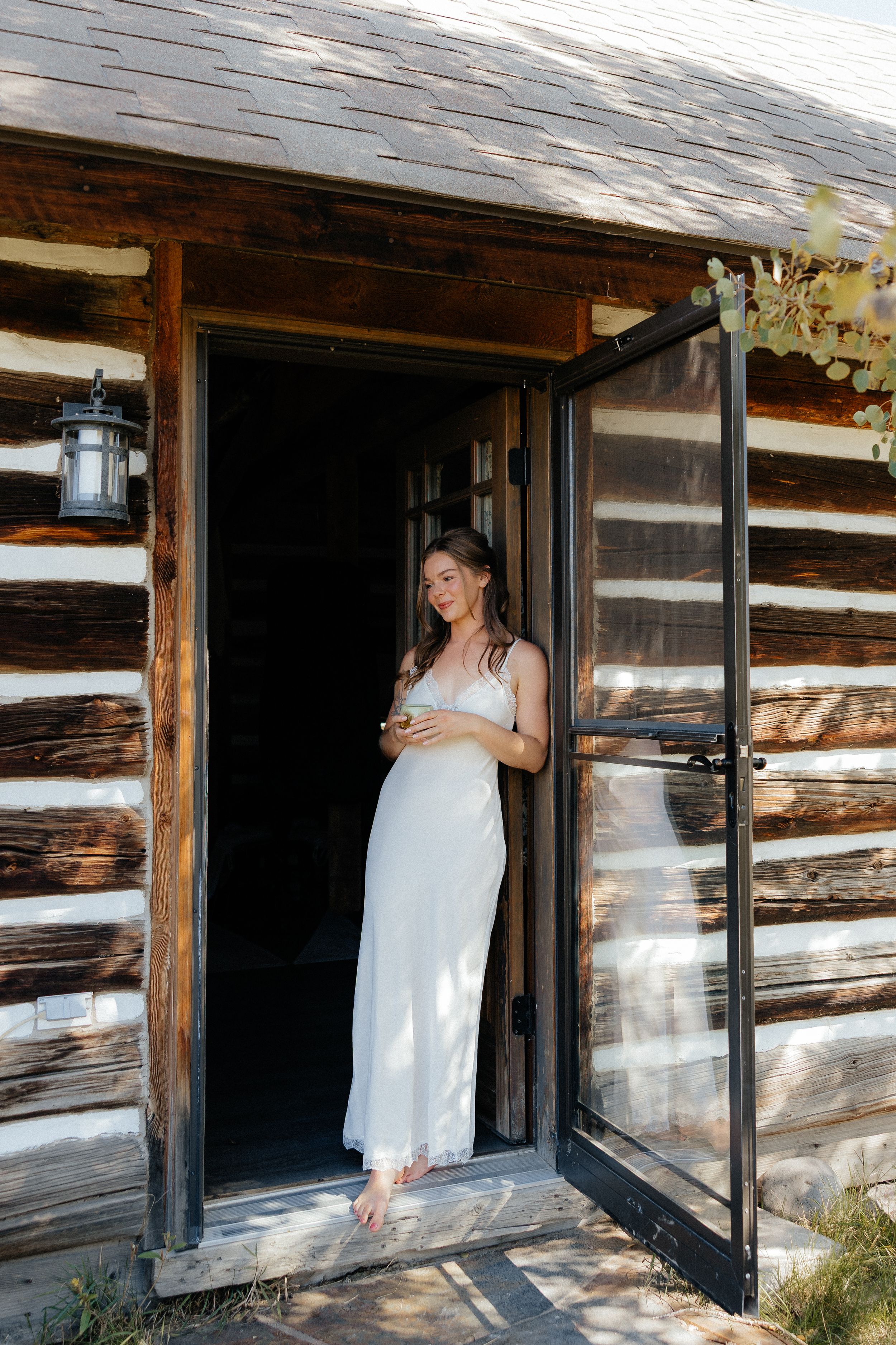 A rustic log cabin entrance with a white dress in the doorway on a sunny day.