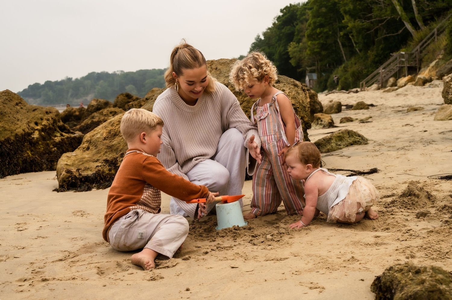 Family enjoys beach time together while playing with bucket on sandy shore near rocky coastline.