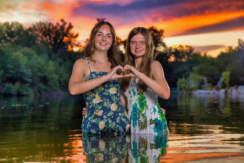 Two people standing in lake water at sunset making heart shape with hands while wearing matching floral dresses.