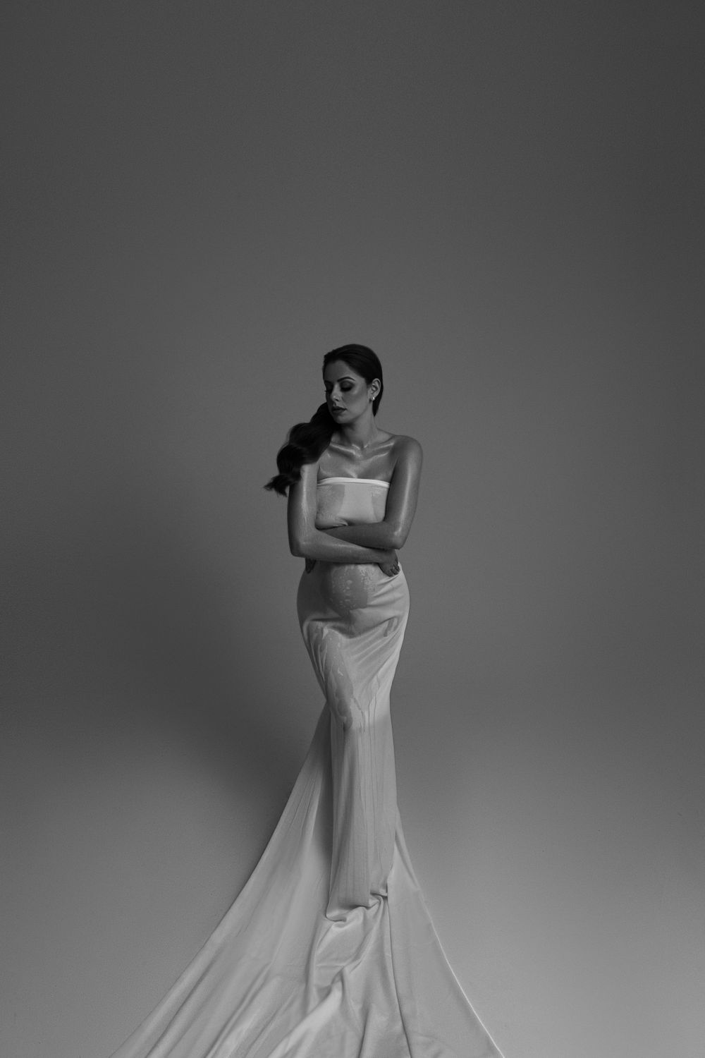 Dramatic black and white maternity photo with flowing white gown against dark background.