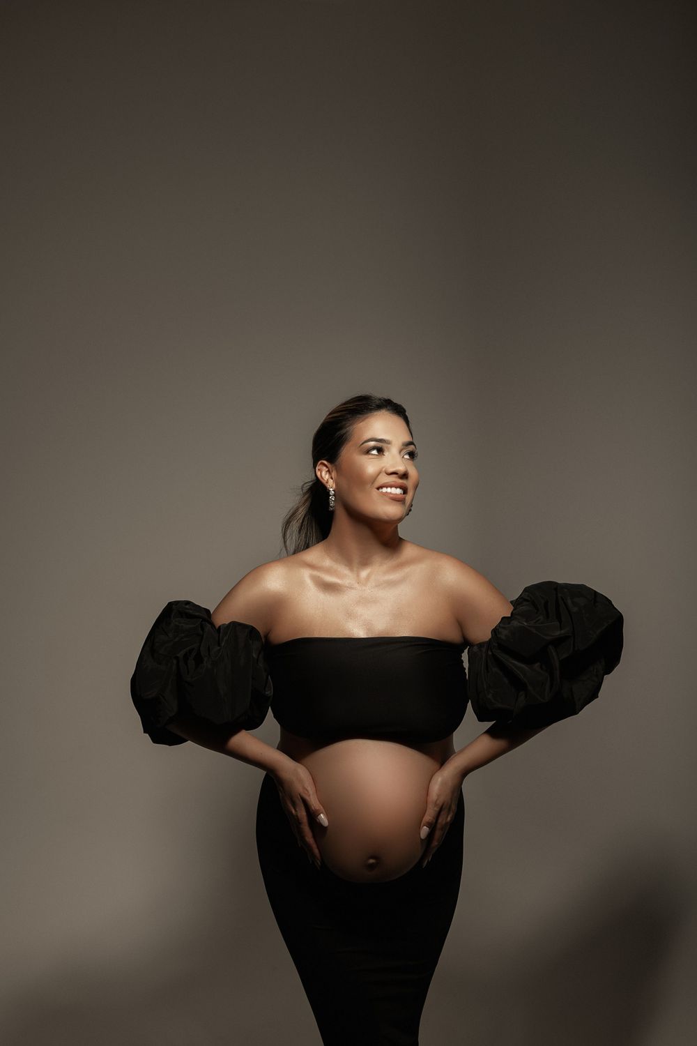 Dramatic studio maternity portrait with black puff-sleeve dress against moody background.