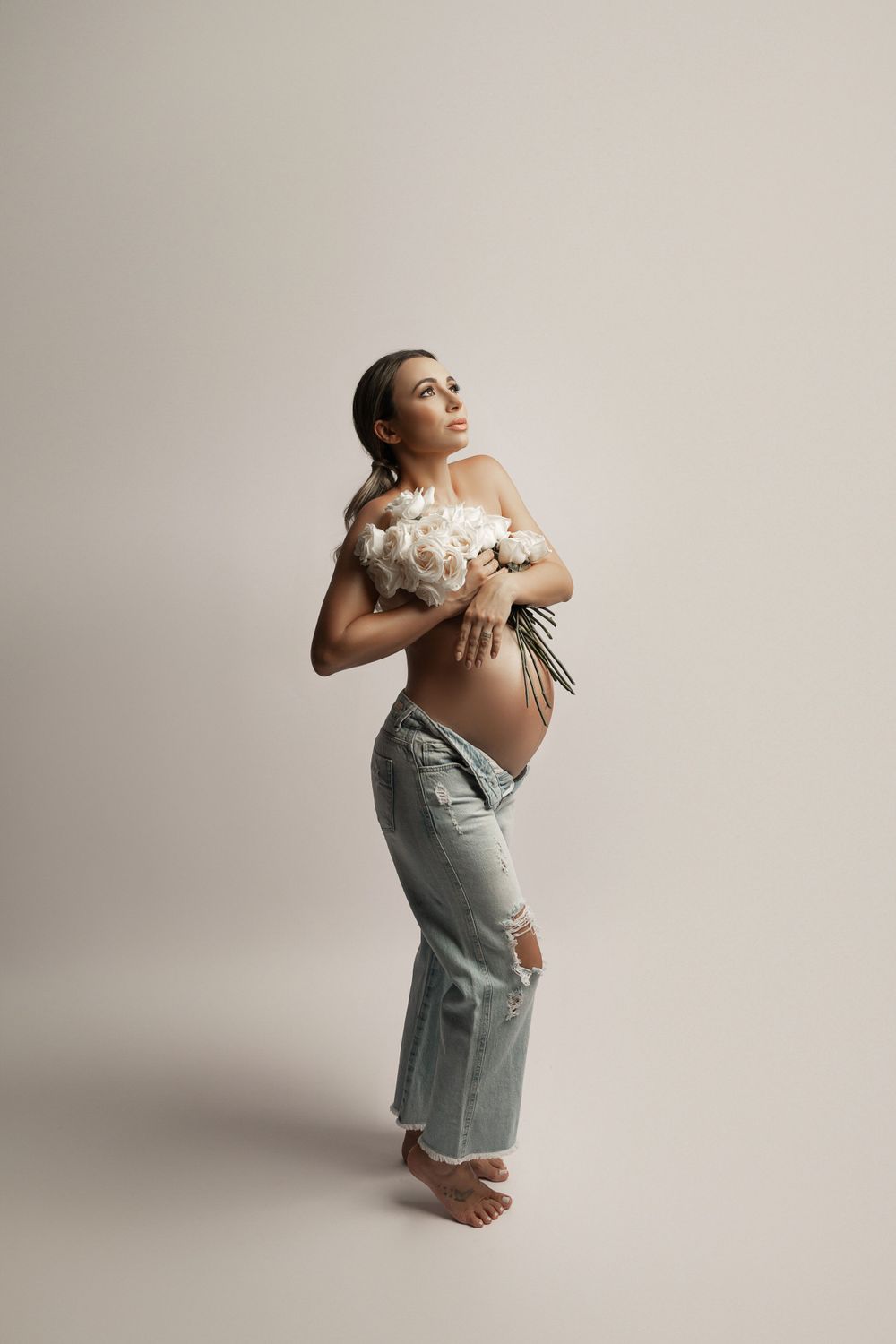 Expectant mother in jeans holding white flowers against neutral backdrop in artistic maternity portrait.