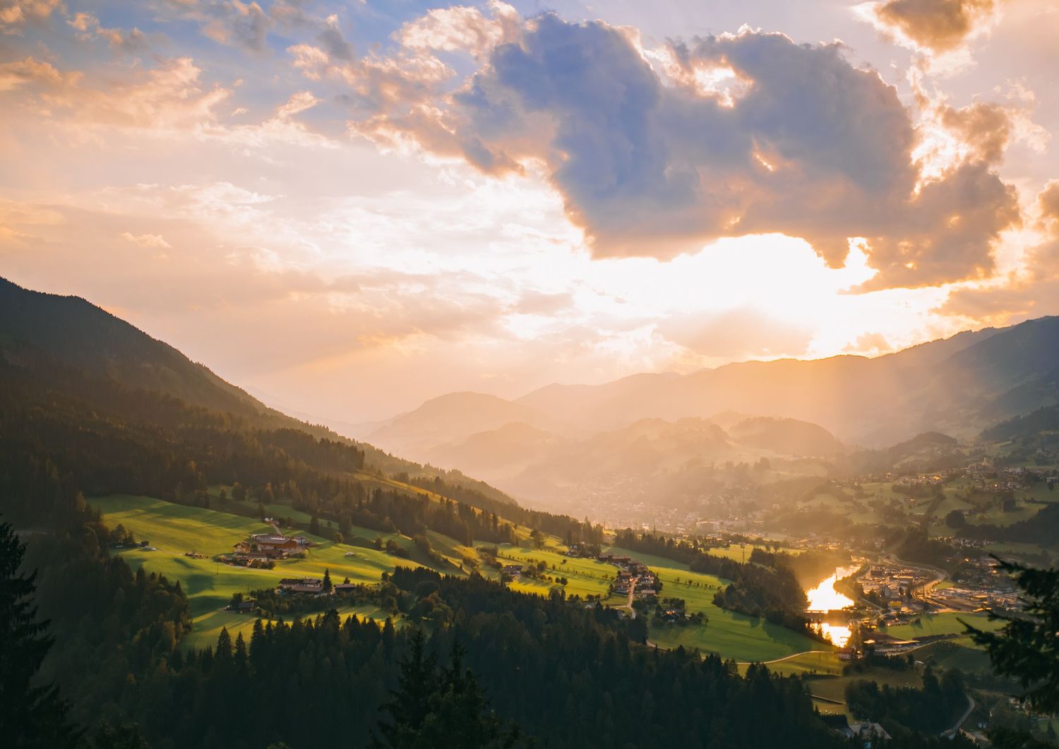 Sunlit mountain valley with green meadows and dramatic clouds during golden hour sunset.
