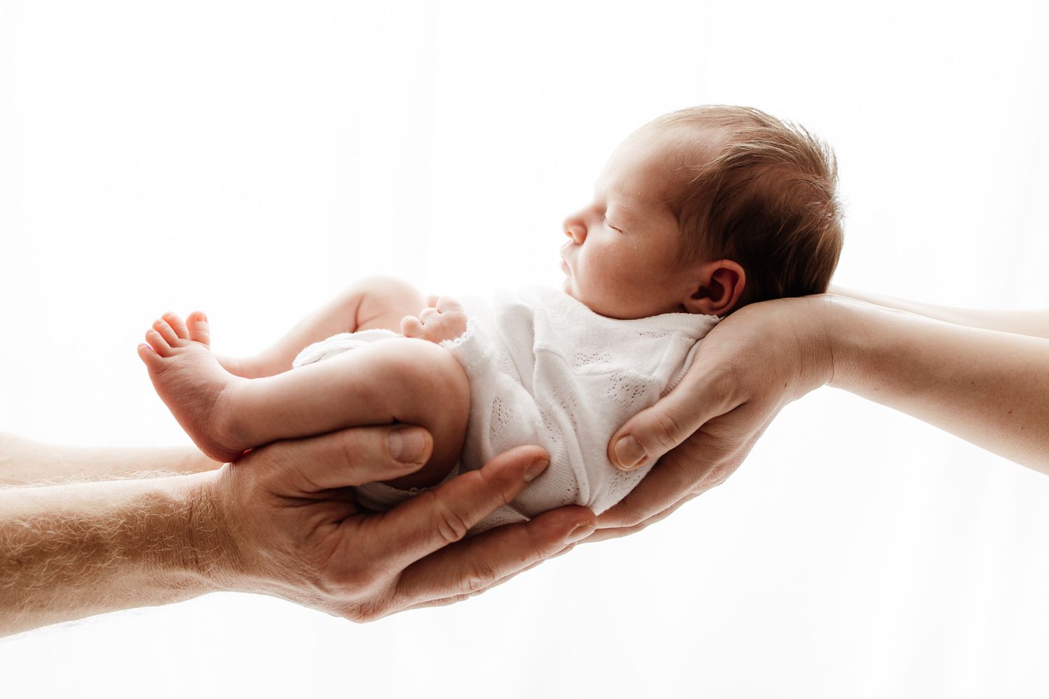 Gentle hands carefully hold a sleeping newborn baby wearing white against a bright background.